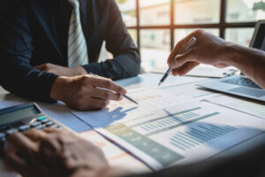 The image shows a business meeting or financial discussion, with professionals analyzing charts, graphs, and financial reports in a well-lit office setting.